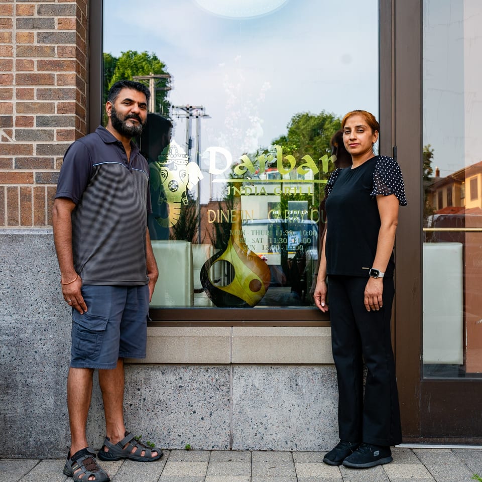 Two people, a man and a woman, stand outside Darbar India Grill & Bar, with its logo visible.