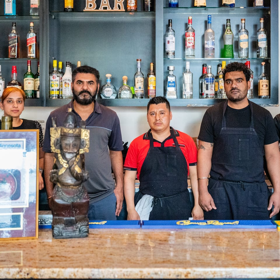 Four Darbar India Grill & Bar staff members stand behind a bar with liquor bottles.