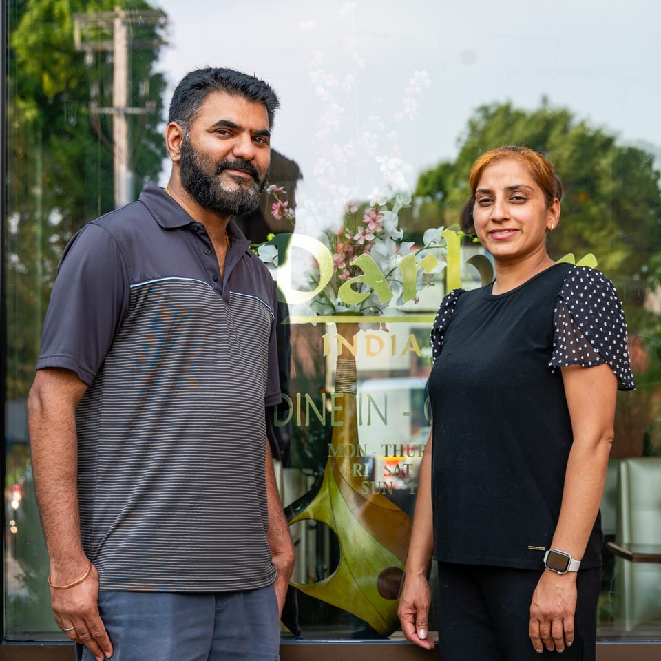 Two Darbar India Grill & Bar owners, a man and a woman, stand smiling in front of the restaurant.